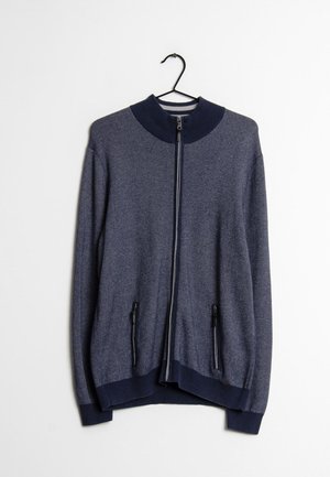 Granatowy sweter na zamek błyskawiczny z dwoma kieszeniami na zamek, wiszący na czarnym wieszaku na białym tle.