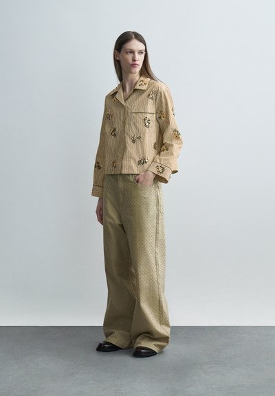 Γυναίκα που στέκεται, φοράει μπλούζα μακριών μανικιών με μπεζ ρίγες και floral μοτίβο και φαρδιά μπεζ παντελόνια με σχέδιο, μαζί με μαύρα παπούτσια, μπροστά σε απλό φόντο.