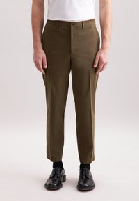 Pantaloni marroni su misura con una vestibilità slim, realizzati in tessuto liscio, con tasche frontali e chiusura a bottone. Indossati con scarpe nere e calzini.