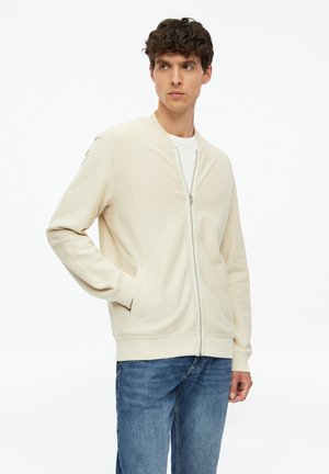 Junger Mann trägt eine cremefarbene Jacke mit Reißverschluss über einem weißen Hemd und blauen Jeans, steht mit einer Hand in der Tasche und schaut zur Seite.