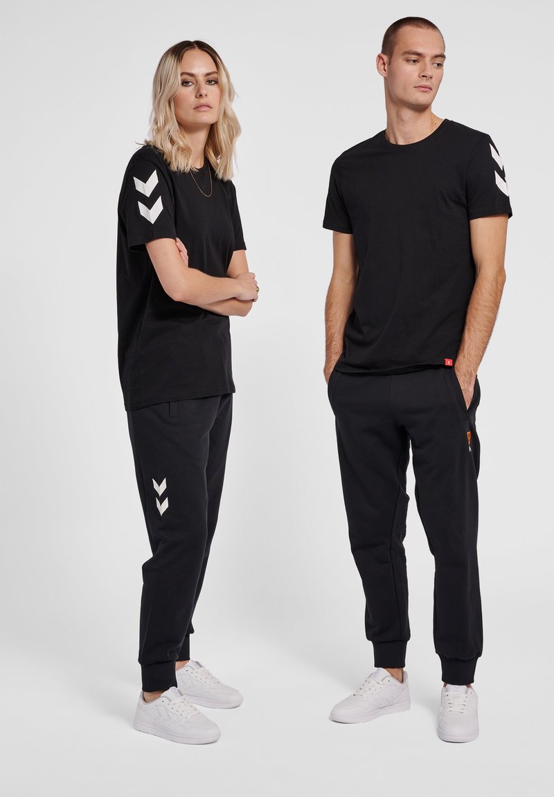 Zwei Erwachsene tragen passende schwarze T-Shirts und Jogginghosen mit weißen Chevron-Logos und stehen vor einem schlichten hellen Hintergrund.
