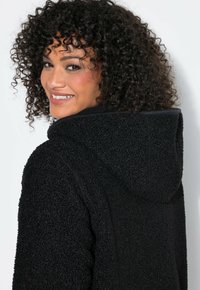 Mujer con cabello rizado girando la cabeza y sonriendo, vistiendo una chaqueta con capucha negra y textura, sobre un fondo claro y liso.
