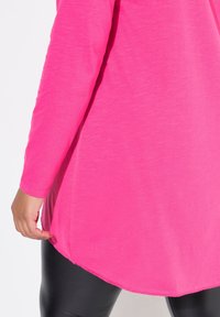 Haut fuchsia à manches longues avec un ourlet incurvé et une texture douce, associé à un legging noir. Design simple, sans motifs ni accessoires visibles.