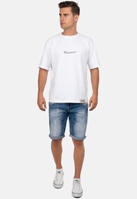 T-shirt en coton blanc avec un logo en script noir, associée à un short en denim bleu clair, à manches retroussées, et des baskets blanches. Design simple et décontracté.