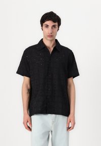Chemise noire à manches courtes avec des motifs brodés, fermeture à boutons à l'avant, et un tissu texturé. Associée à un pantalon en denim de couleur claire.