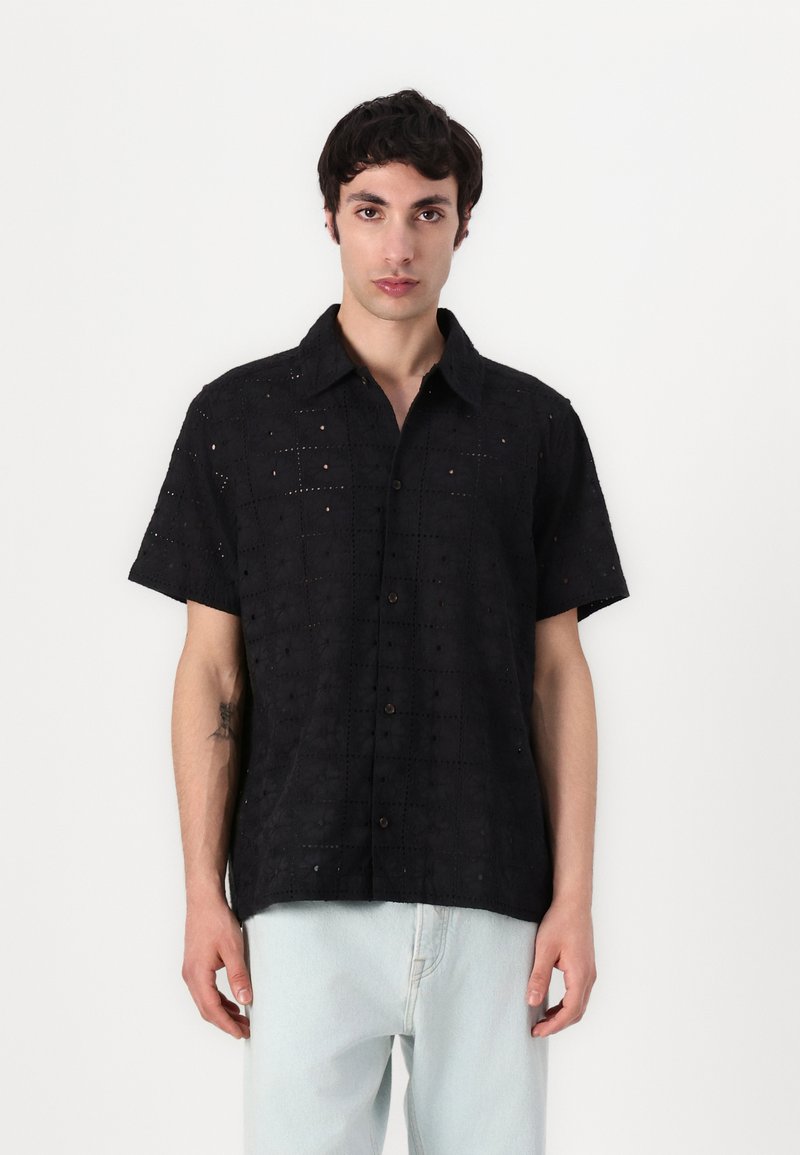 Camisa preta de mangas curtas com padrões bordados, frente abotoada e tecido texturizado. Combinada com calças de ganga de tom claro.