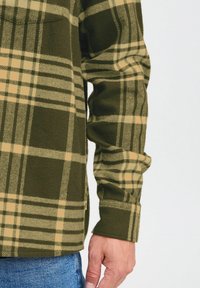 Rude fleece skjorte, olivengrøn med beige striber, har lange ærmer og en sideslids fra kanten. Blød tekstur med synlige sømdetaljer.