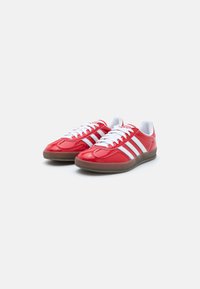 Red synthetic sports shoes with glossy finish, white stripes, and lace closure. Features a rubber sole and rounded toe design.