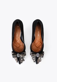 Ballerines en suede noir avec une semelle intérieure matelassée marron, ornées d'un grand nœud décoré de strass noirs, dorés et transparents à l'avant.