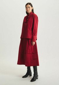 Robe rouge à couches avec un col froncé, des manches longues et une silhouette fluide, associée à des bottes noires à talons montantes.