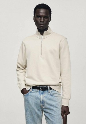 Helle beige Zip-Up Sweatshirt mit gerippter Textur und einem Viertelreißverschluss-Kragen. Getragen mit verblassten blauen Jeans und einem schwarzen Gürtel.