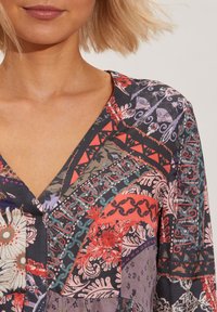 Kvinna i en flerfärgad lappad V-ringad topp med blommiga och geometriska mönster, kort blont hår och naturlig sminkning.