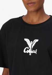 Zwarte katoenen T-shirt met een prominent wit grafisch ontwerp van een engel met vleugels en de tekst "Colucci" eronder. Ronde halslijn, losse pasvorm.