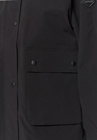 Zwarte jas met een voorzak met klep en drukknoopjes, gemaakt van textuurstof, met een strakke, minimalistische uitstraling.