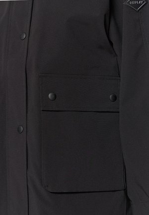 Schwarze Jacke mit einer vorderen Tasche mit Klappe und Druckknöpfen, aus strukturiertem Stoff gefertigt, zeigt ein elegantes, minimalistisches Design.