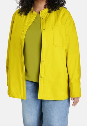 Geel knopenjasje met een kraag, voorzak en drukknoopsluiting, gelaagd over een groene shirt en gedragen met een blauwe spijkerbroek.