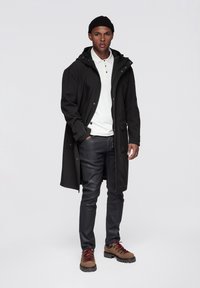 Czarny długi płaszcz na białej koszulce polo, zestawiony z ciemnymi spodniami i brązowymi butami trekkingowymi z czerwonymi sznurówkami. Model stoi na tle jednolitego tła.