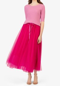 Žena ve vínové žebrované blůze s rukávy do lokte, jasně růžové midi sukni z tylu, která je svázaná stuhou, a tělových sandálech na kotníku.