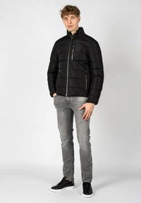 Giubbotto imbottito nero con chiusura a zip, cuciture verticali e due tasche laterali. Indossato sopra jeans grigi e sneaker nere, con colletto.