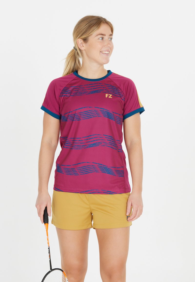 Leende kvinna i träningskläder som håller en badmintonracket, klädd i en magenta och marinblå tröja med senapsgula shorts, ser åt sidan.