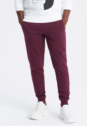 Bordeaux joggerbukser lavet af blødt stof, med sidelommer, tætsiddende design og ribbede ankelmanchetter. Sat sammen med hvide sneakers.