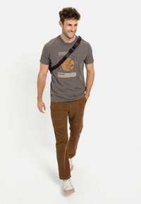 Graues Grafik-T-Shirt mit einem Bergdesign, kombiniert mit braunen Cordhosen und weißen Turnschuhen. Eine schwarze Umhängetasche wird getragen.