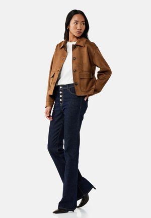 Kvinde iført brun jakke med knapper, hvid skjorte, mørke højttaljede jeans med synlige knapper og sorte hælede sko, står med den ene hånd i lommen.