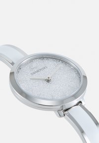 Orologio Swarovski con cassa rotonda in tonalità argento, cinturino in ceramica bianca e quadrante argentato con una texture simile a cristallo. Presenta lancette e indicatori argentati.