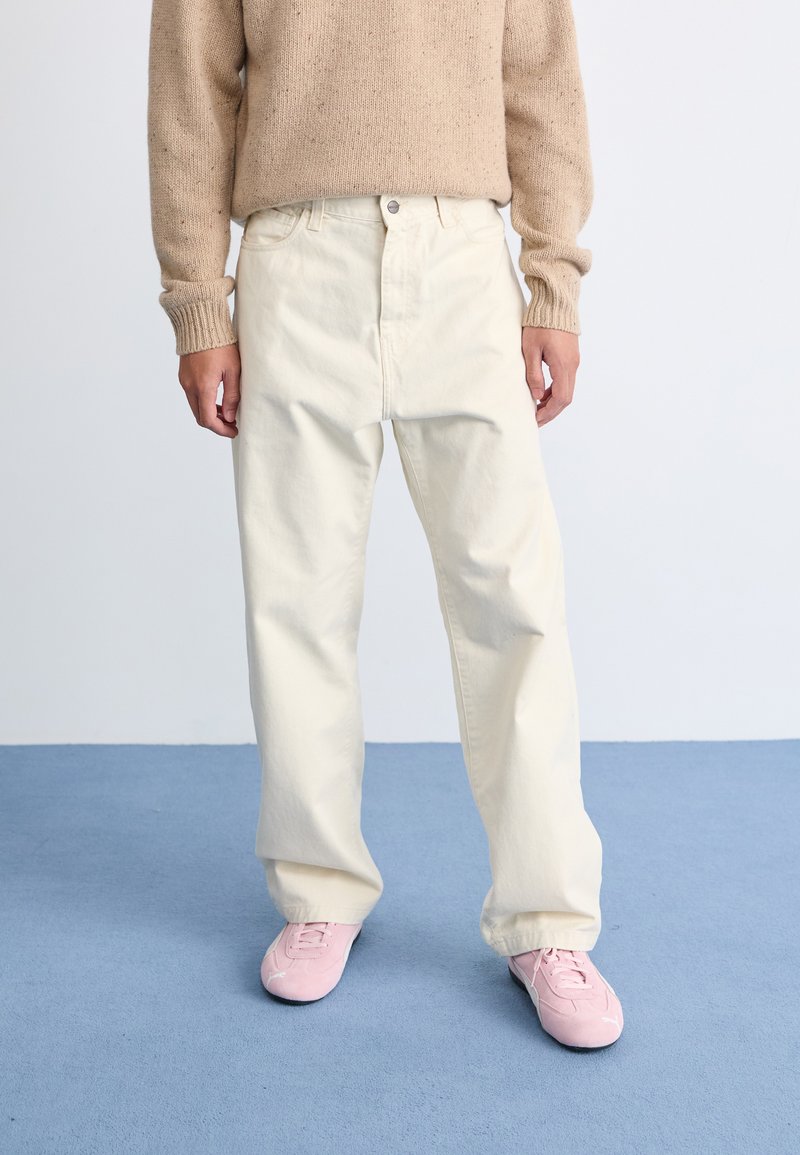 Maglione lavorato a maglia beige, pantaloni larghi di colore bianco sporco e sneakers rosa chiaro. I pantaloni hanno tasche anteriori e chiusura con bottone.