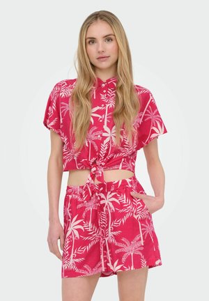 Jeune femme portant un haut rouge à manches courtes avec imprimé tropical, boutonné et noué à la taille, ainsi qu'un short assorti, debout avec une main dans la poche.
