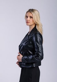 Blond kvinna i svart läderjacka och svart klänning, poserande på sidan med händerna hållande dragkedjan på jackan mot en enkel ljus bakgrund.