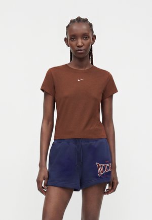 Γυναίκα με πλεξούδες φοράει καφέ μπλουζάκι Nike και σκούρο μπλε σορτς Nike, στέκεται μπροστά σε απλό φωτεινό φόντο.