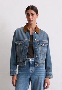 Veste en denim avec un col en velours côtelé marron, comportant des poches avant, des boutons et un délavage bleu moyen. Portée avec un bas en denim assorti.