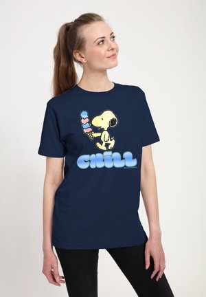 Jonge vrouw draagt een marineblauw T-shirt met Snoopy die een hoorntje met drie bolletjes ijs vasthoudt en het woord "Chill" in blauwe bubbelletters.