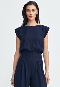 Granatowa bluzka z rękawami w kształcie rękawiczek, z marszczoną talią i gładką fakturą, w zestawieniu z pasującymi wysokimi spodniami z plisami.