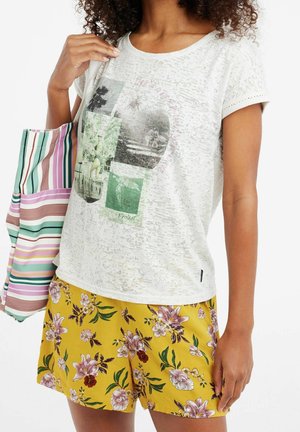 Frau trägt ein weißes T-Shirt mit Grafik und gelbe florale Shorts und hält eine gestreifte Tragetasche über der Schulter.