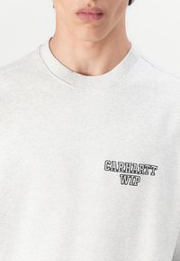 Grå sweatshirt lavet af bomuld, med ribbet rund hals og et lille, sort broderet logo på venstre bryst. Glat tekstur.
