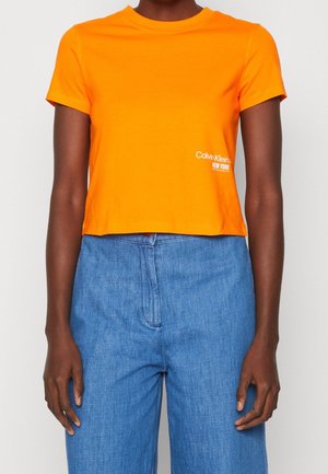 Osoba v jasně oranžovém krátkém tričku crop top s malým bílým logem Calvin Klein a v modrých džínech s vysokým pasem.