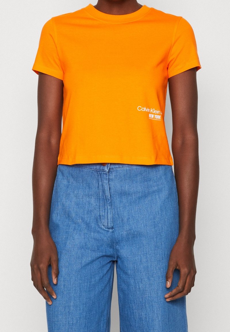 Henkilö pukeutuneena kirkkaan oranssiin lyhythihaiseen crop-toppiin, jossa on pieni valkoinen Calvin Klein -logo, ja korkeavyötäröisiin sinisiin farkkuihin.