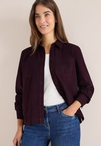 Bordeaux knap-ud skjorte med to forlommer, lavet af tekstureret stof. Matchet med en hvid T-shirt og blå denim jeans.