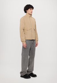 Beige jakke med høy krage, trykknapper og to store frontlommer, sammen med grå jeans og svarte sko. Glatt tekstur, uformelt design.