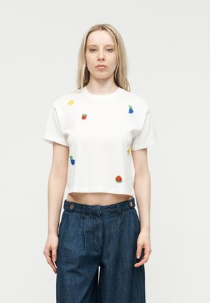 Jeune femme aux longs cheveux blonds portant un crop top blanc orné de petites broderies de fruits colorés et un pantalon en denim bleu taille haute.