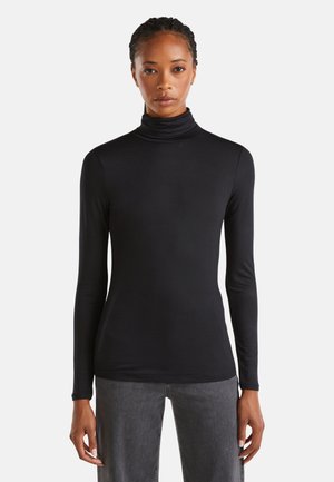 Vrouw in een aansluitend zwart coltrui met lange mouwen en grijze broek, staand tegen een witte achtergrond.