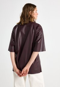 Mørkerød faux læder top med korte ærmer, glat tekstur, løs pasform og minimale sømme, parret med hvide bukser, vist fra ryggen.