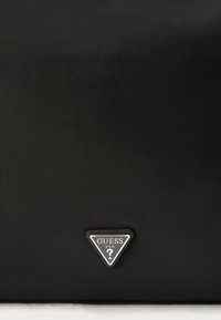 Μαύρη υφασμάτινη τσάντα tote με τρίγωνο μεταλλικό λογότυπο που αναγράφει "GUESS" και ένα ερωτηματικό. Μινιμαλιστικός σχεδιασμός χωρίς επιπλέον μοτίβα.