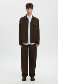 Ruda didelio dydžio striukė su kišenėmis, dėvima ant šviesiai mėlynos dryžuotos marškinių, derinama su laisvomis rudomis kelnėmis ir juodais batais.