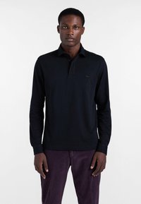 Schwarzes langärmliges Poloshirt mit Kragen, das ein dezentes Logo auf der Brust trägt. Kombiniert mit lila Cordhosen.