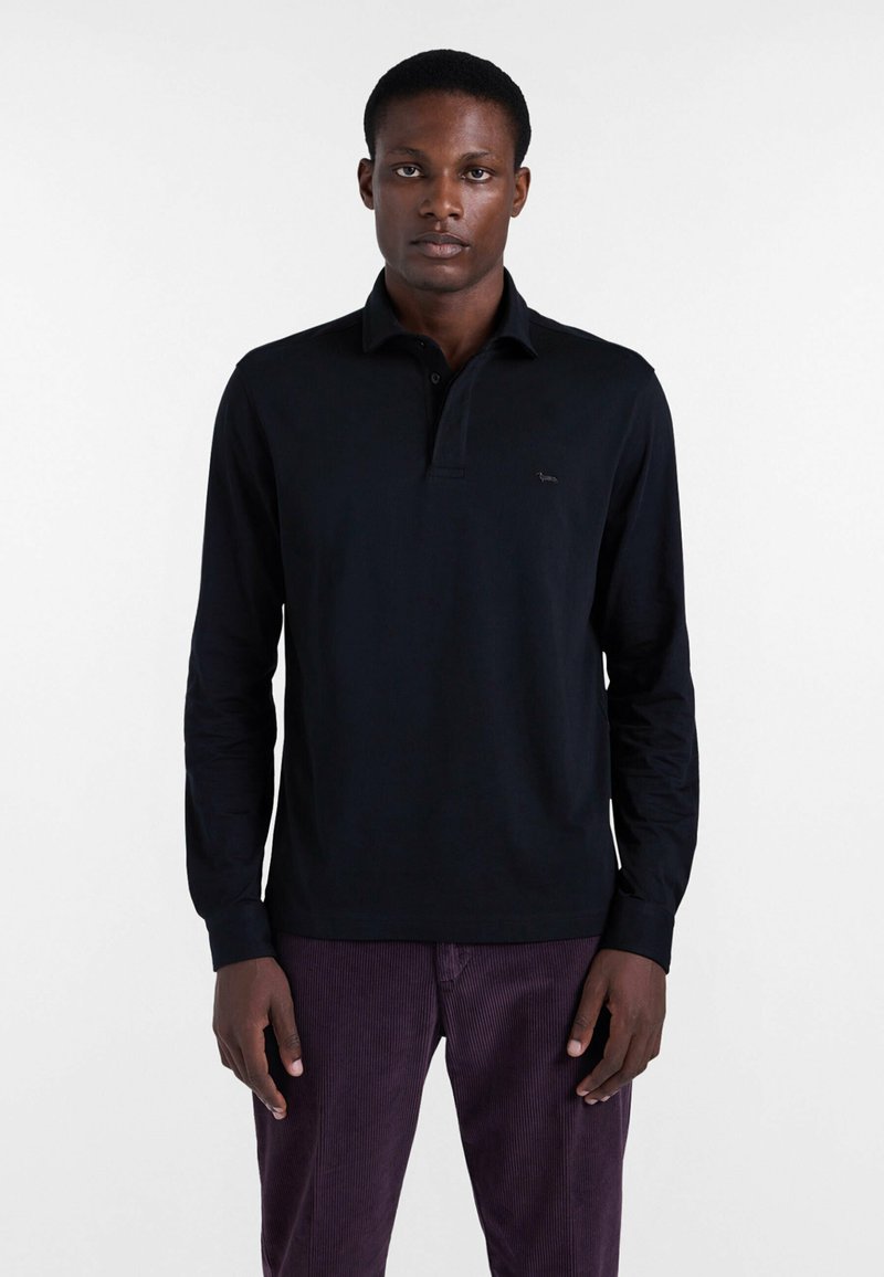 Schwarzes langärmliges Poloshirt mit Kragen, das ein dezentes Logo auf der Brust trägt. Kombiniert mit lila Cordhosen.