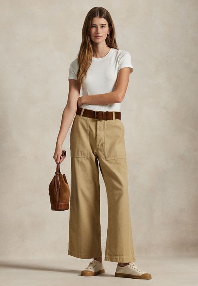 Γυναίκα που στέκεται, φορώντας λευκό t-shirt, μπεζ παντελόνι φαρδιάς γραμμής με καφέ ζώνη, λευκά αθλητικά παπούτσια, κρατώντας καφέ σουέντ τσάντα κουβά από το λουρί.