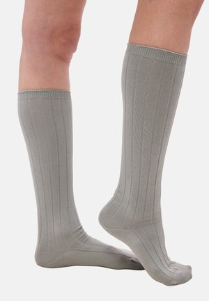 Beine mit knielangen, hellgrauen, gerippten Socken, ein Fuß flach auf dem Boden, der andere leicht auf den Spitzen gehoben.